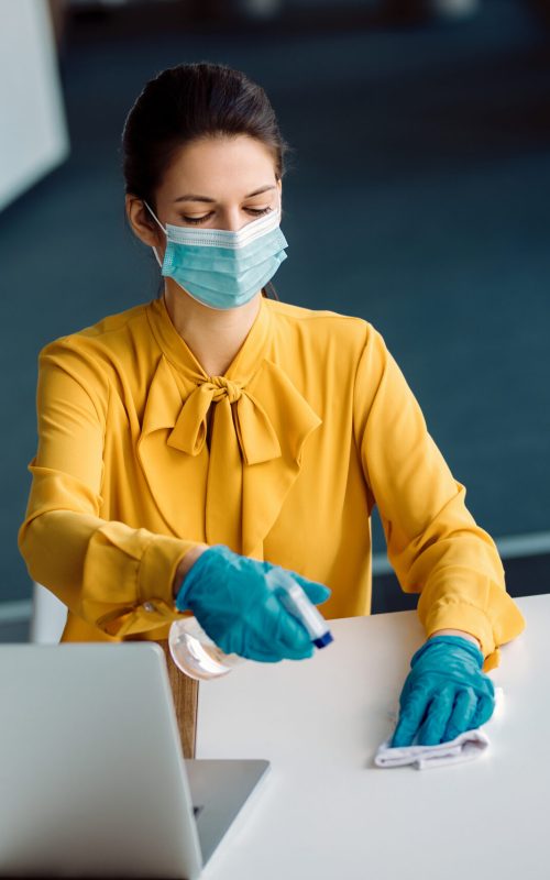 Female entrepreneur wearing face mask and gloves while cleaning her desk with disinfectant in the office.